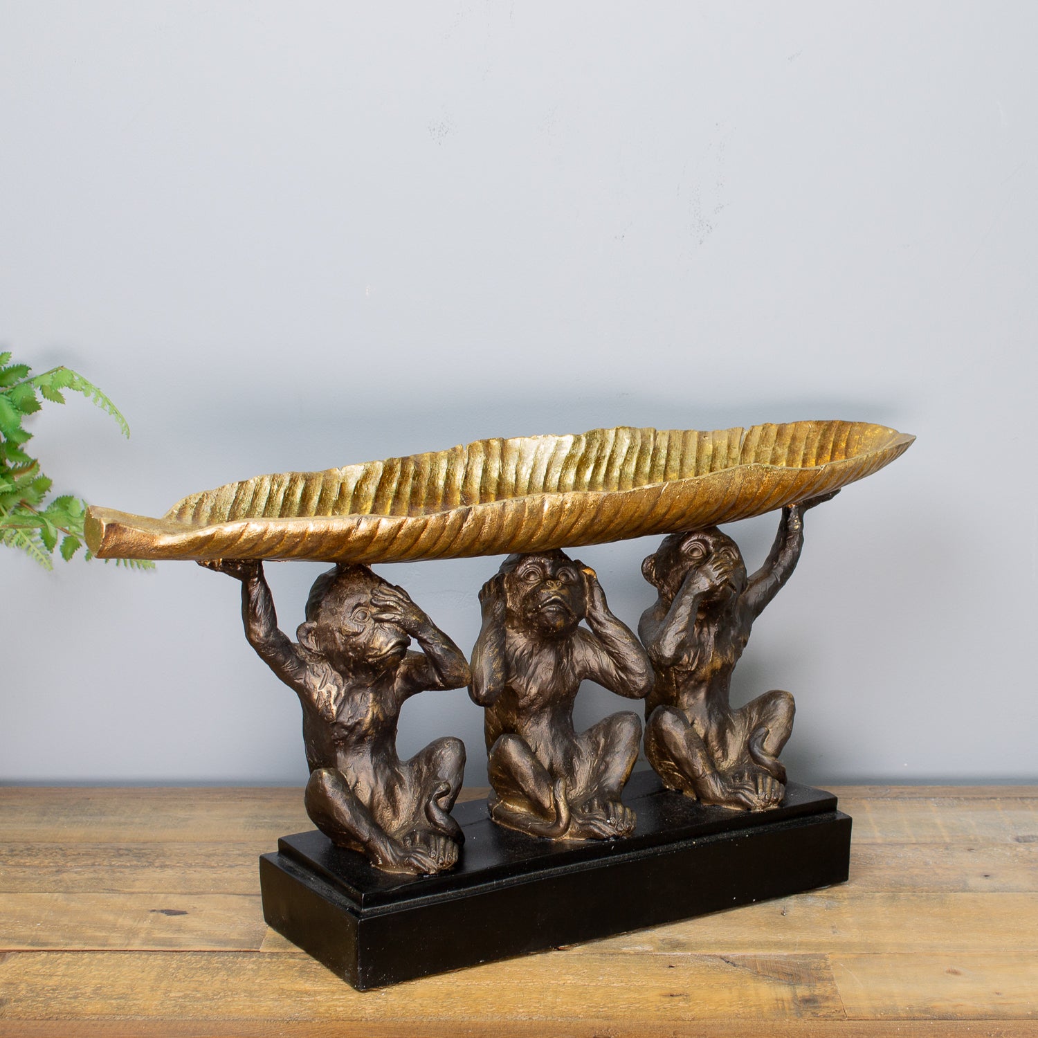 Decorative sculpture of three monkeys holding a large shell on a wooden surface with a gray background.