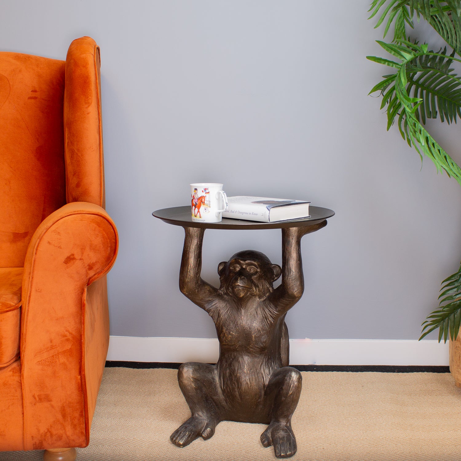 Bronze monkey table with a cup and book next to an orange armchair against a gray wall.