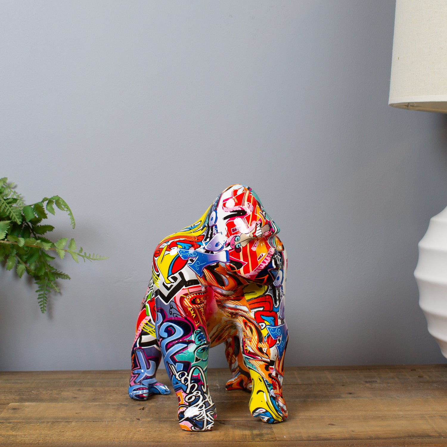 Colorful gorilla statue on a wooden surface with a gray wall and plant in the background